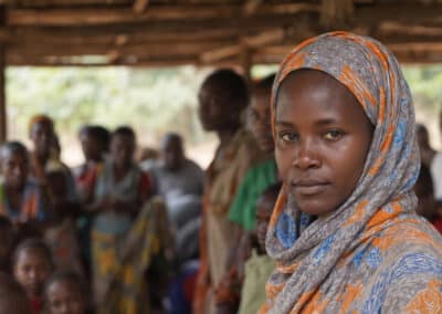 Woman wearing colorful headscarf prominent in group at village gathering Focus on serious expression looking towards camera blurred background of African community Represents culture tradition